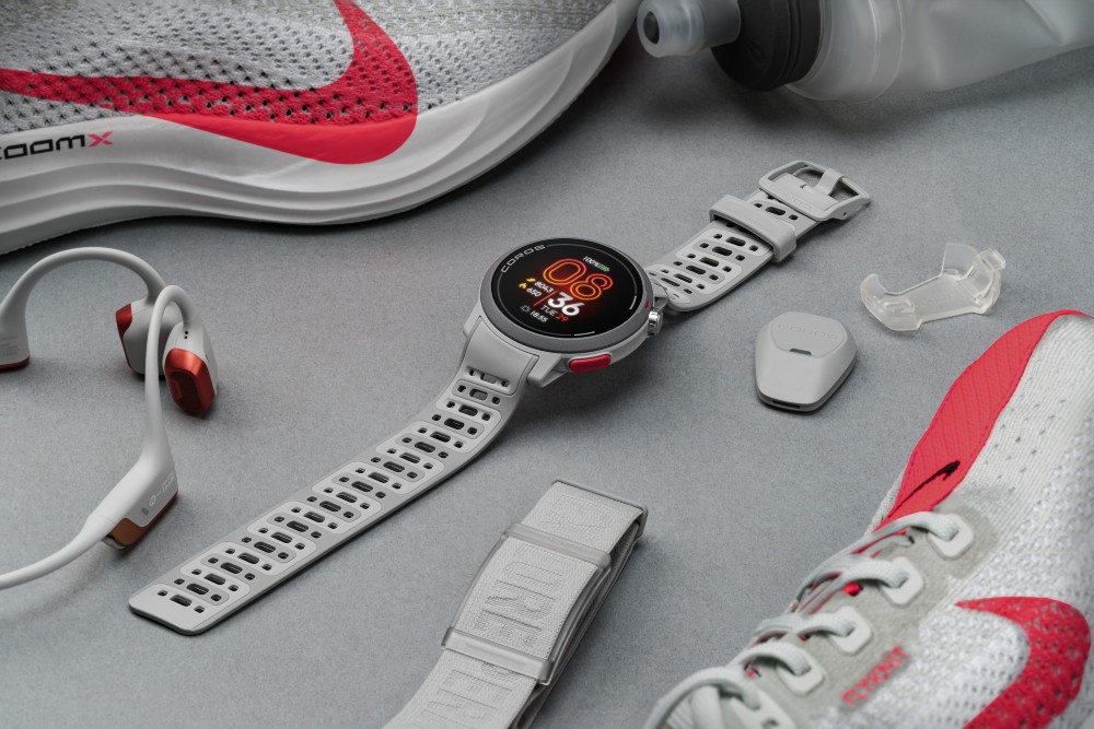 A grey Coros Pace Pro running watch surrounded by running shoes, some headphones and a water bottle.